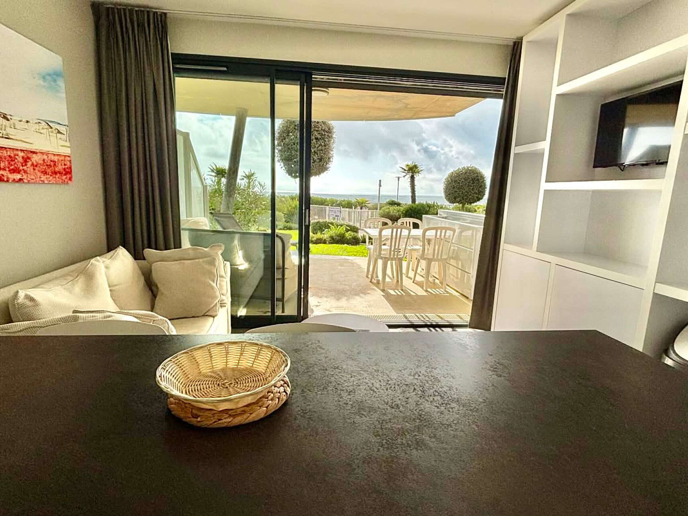 A room with a dark table, a wicker basket, a sofa with cushions, a TV on a shelf, and a view of the outdoors through a sliding door.