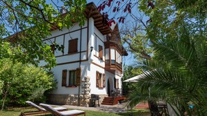 Ein zweistöckiges Haus mit einem Balkon, einer kleinen Terrasse mit Tisch und Stühlen sowie einem Garten mit einer Palme.