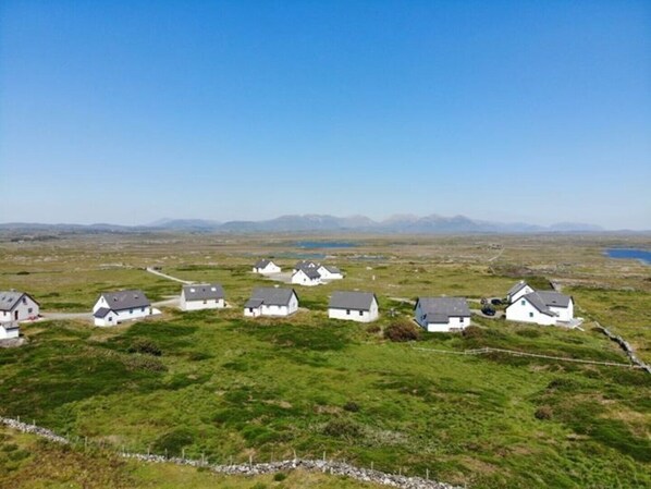 A cluster of white houses with grey roofs on a grassy hill, surrounded by open fields and distant mountains.