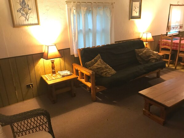 A cozy living room with a dark green sofa, two wooden side tables with lamps, a wooden chair, and a small wooden table. - Budget Rustic Family Cabin (Wilmington)