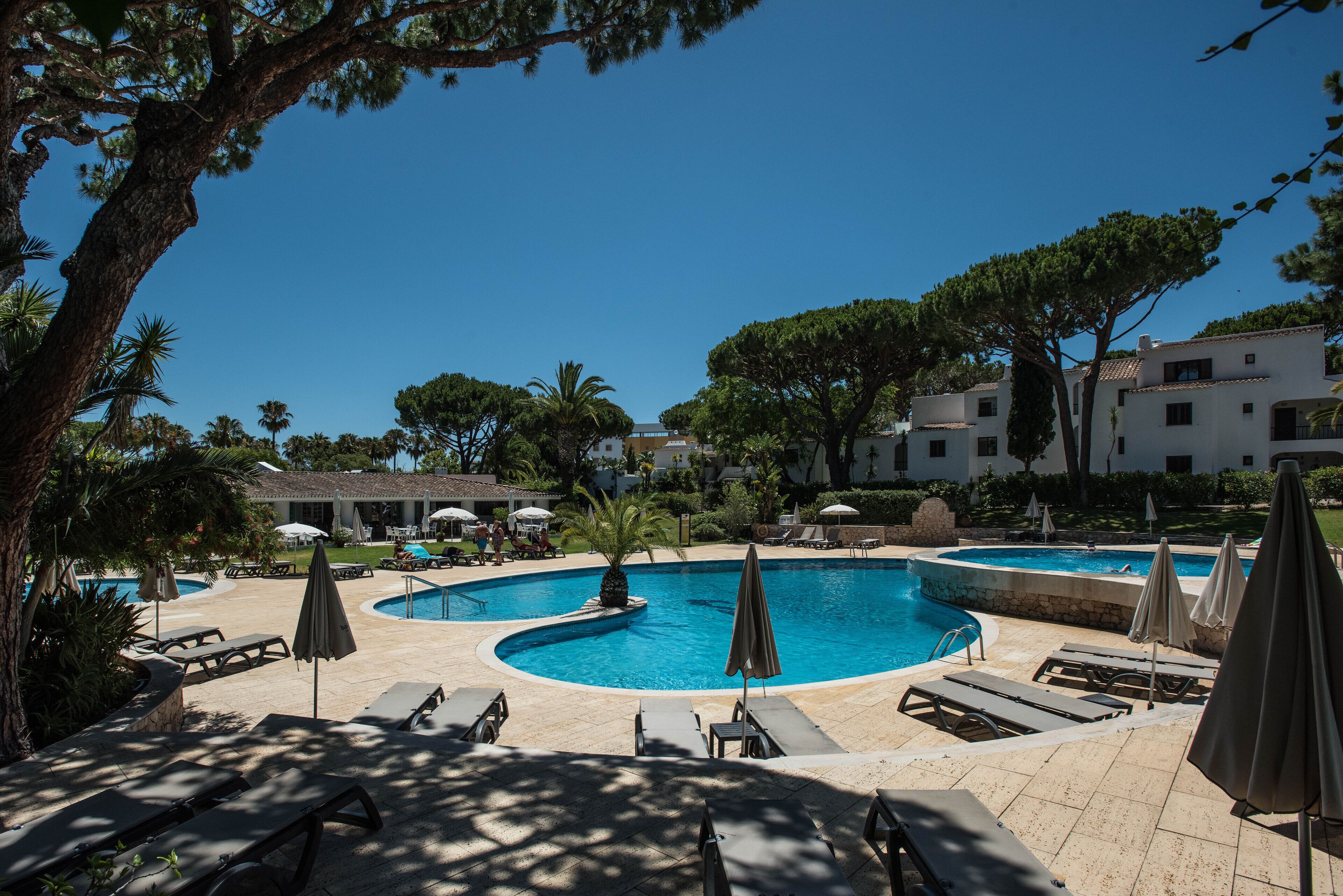A swimming pool area with lounge chairs and umbrellas, surrounded by trees and buildings.