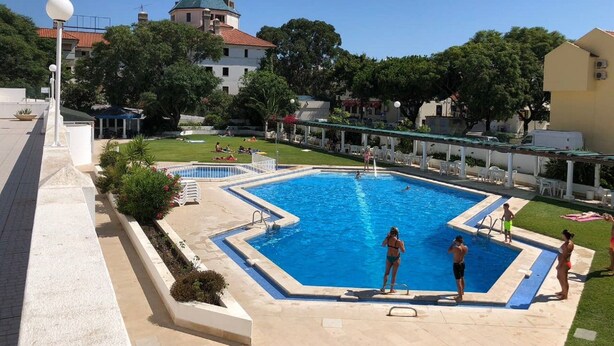Una piscina con un área pequeña para buceo, rodeada por un solárium blanco, con un edificio y árboles al fondo.