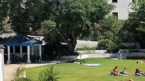 Zona de piscina con área de descanso cubierta, césped y un edificio al fondo.