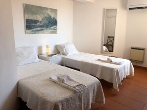 Two single beds with white bedspreads and towels, a nightstand with a lamp and book, and a large wave painting adorn this cozy space.