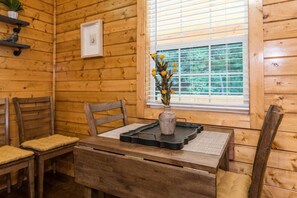 A wooden cabin interior with a dining table, two chairs, a vase with yellow flowers, and a window with blinds.