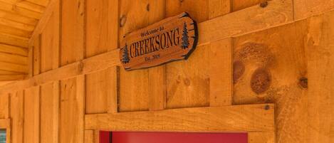Un mur extérieur en bois, avec une porte rouge et une enseigne sur laquelle on peut lire : « Bienvenue chez CREEKSONG, fondé en 2019 ».