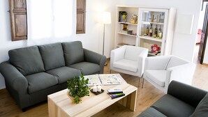 Un salón con un sofá gris, un sillón blanco, una mesa de centro de madera con una planta y libros, y una estantería con diversos objetos.