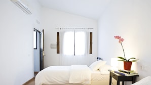 Un dormitorio con cama, una mesita de noche con libros, una planta en maceta y una ventana con cortinas.