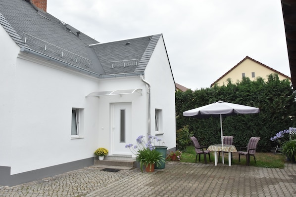 A white house with a grey roof, a small front porch, and a patio area with a table and chairs.