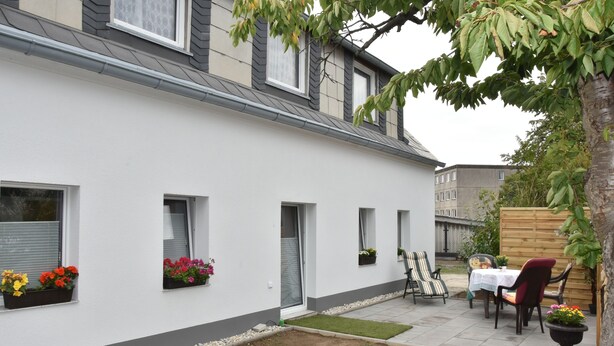 A white building with multiple windows, a small garden area with a table and chairs, and a wooden fence in the background. - King cottage in the center (Königsbrück)