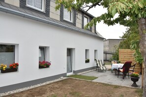 Un edificio blanco con varias ventanas, un pequeño jardín con mesa y sillas, y una valla de madera al fondo.