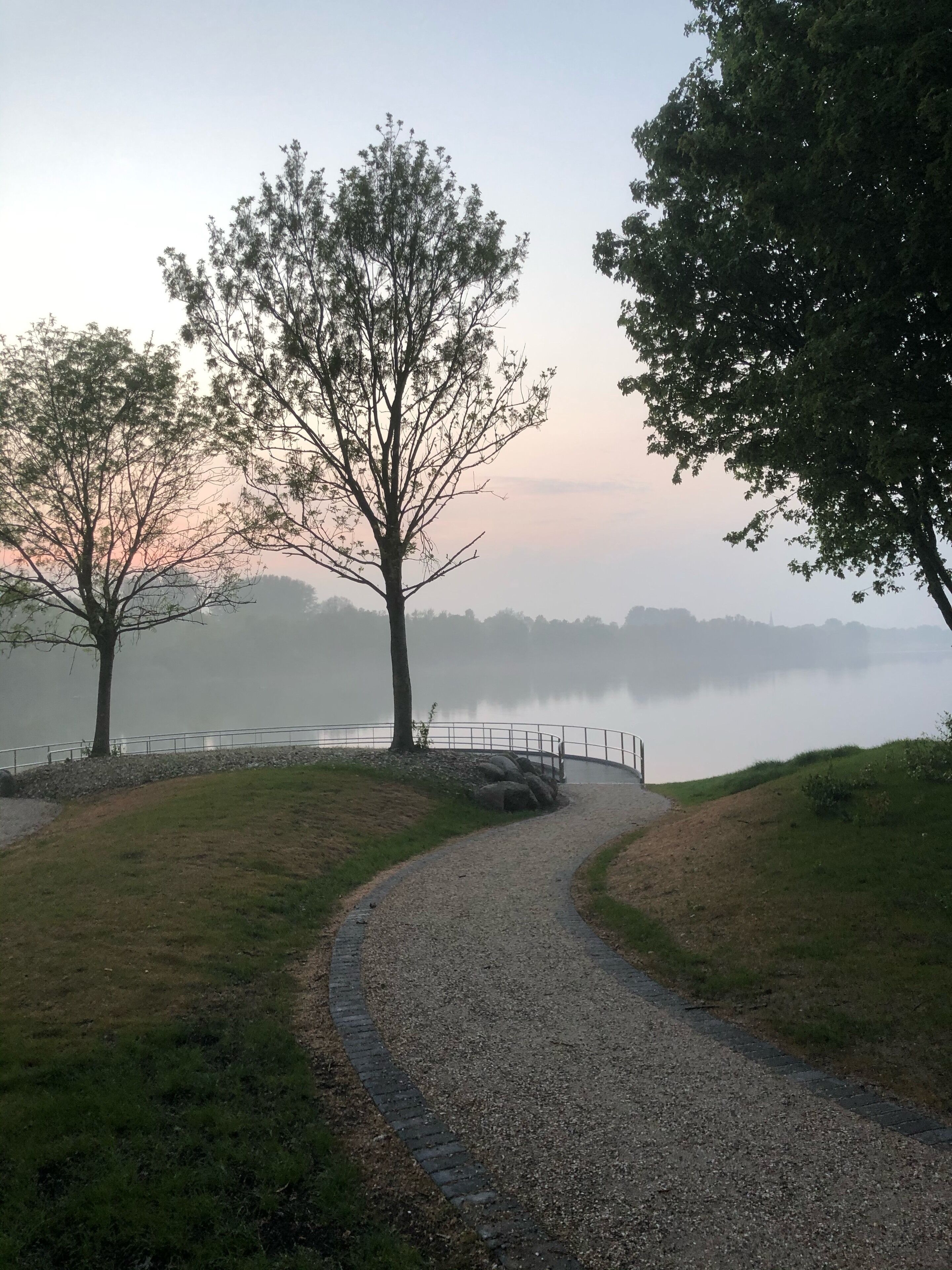 Un sendero de grava serpentea por un parque con árboles y un lago al fondo.