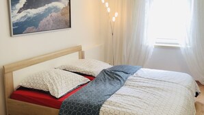 A bed with a wooden frame, a red and white patterned bedspread, and a blue and white blanket. There is a painting of a coastal landscape on the wall above the bed. - Marschenhof at the lakes. (Xanten)
