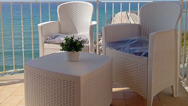 A balcony with two wicker chairs and a small table, overlooking a beach with clear blue water. - Window to the Sea by Alexander 1p (Balestrate)