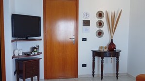 A living room with a television on a stand, a wooden door, a small table with decorative items, and an orange sofa. - Window to the Sea by Alexander 1p (Balestrate)