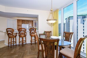 Uma área de refeições com cadeiras de madeira e uma mesa de vidro, uma cozinha com armários de madeira e uma janela grande com vista para edifícios.