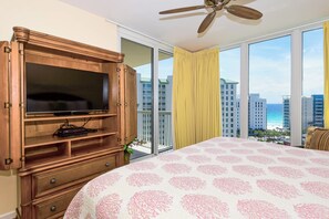 Habitación de hotel con una cama grande, un mueble de televisión de madera, ventilador de techo y balcón con vista a la ciudad.