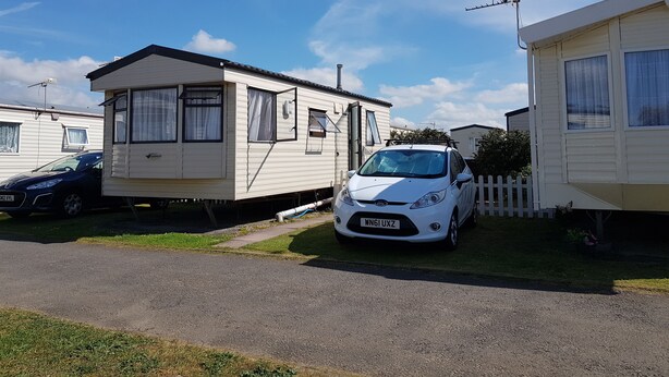 Un terrain de camping avec des mobil-homes, une voiture blanche garée devant l’un d’eux, et un ciel bleu dégagé.