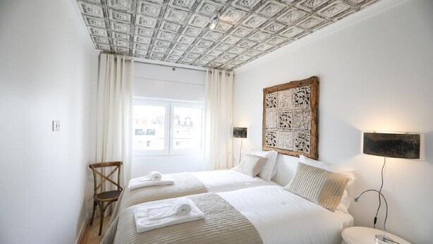 Habitación de hotel con dos camas, cabecero de madera, una mesita de noche con lámpara y una ventana con cortinas.