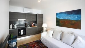 Una cocina moderna con armarios oscuros, horno empotrado y cafetera. En la zona de estar, hay un sofá blanco con cojines y una alfombra colorida. Un cuadro de un paisaje adorna la pared.