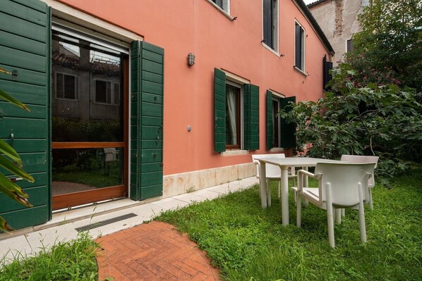 A building with green shutters, a brick pathway, and a table with chairs in the foreground.
