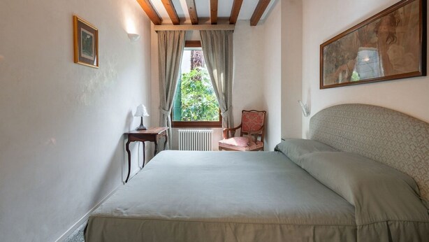 A bedroom with a bed, a chair, a table, and a window with curtains. - Garden Apartment (Venice)