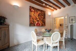 A dining area with a glass table surrounded by white chairs, a large fruit painting on the wall, and a wooden ceiling.
