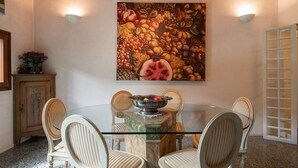 A dining area with a glass table surrounded by chairs, a fruit-themed painting on the wall, and a wooden cabinet with a plant. - Garden Apartment (Venice)