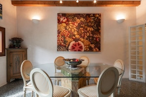 Een eethoek met een glazen tafel, omringd door stoelen, een schilderij met fruitmotieven aan de muur en een houten kast met een plant.