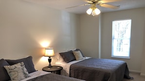 A bedroom with two beds, a nightstand with a lamp, a ceiling fan, and a window with blinds.
