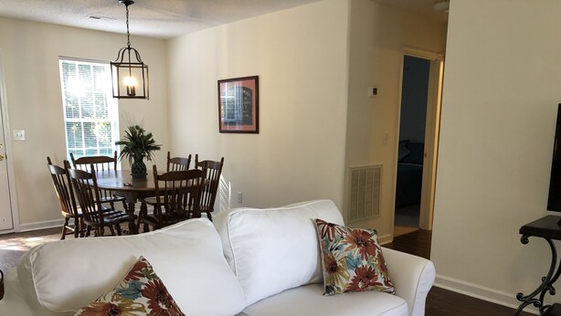 A living room with a white sofa, floral cushions, a dining table with chairs, and a framed picture on the wall.