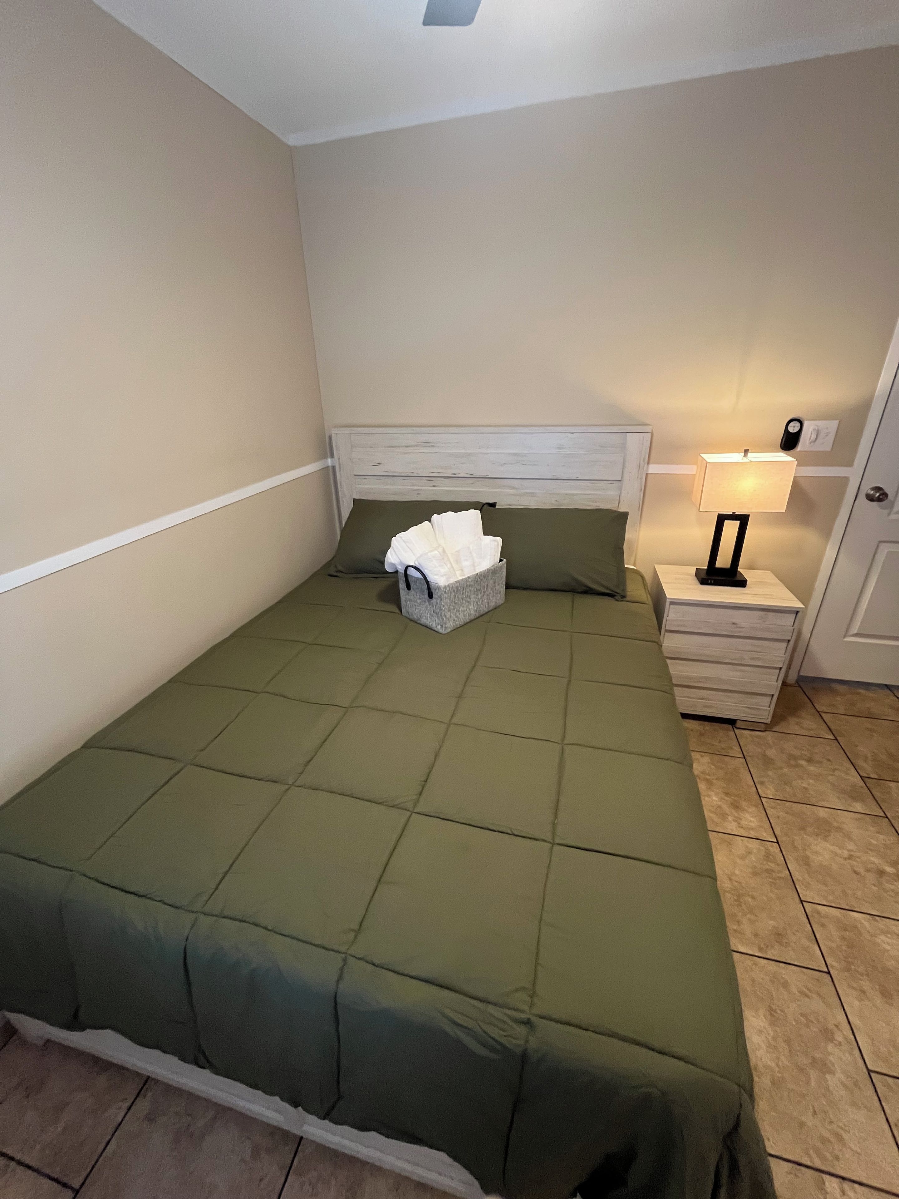 Un letto singolo con una coperta verde, un comodino con una lampada e un cesto di asciugamani bianchi.
