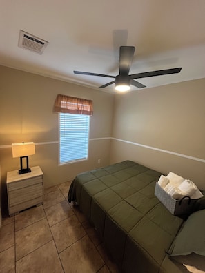 A bedroom with a bed, a nightstand with a lamp, a ceiling fan, and a window with blinds.