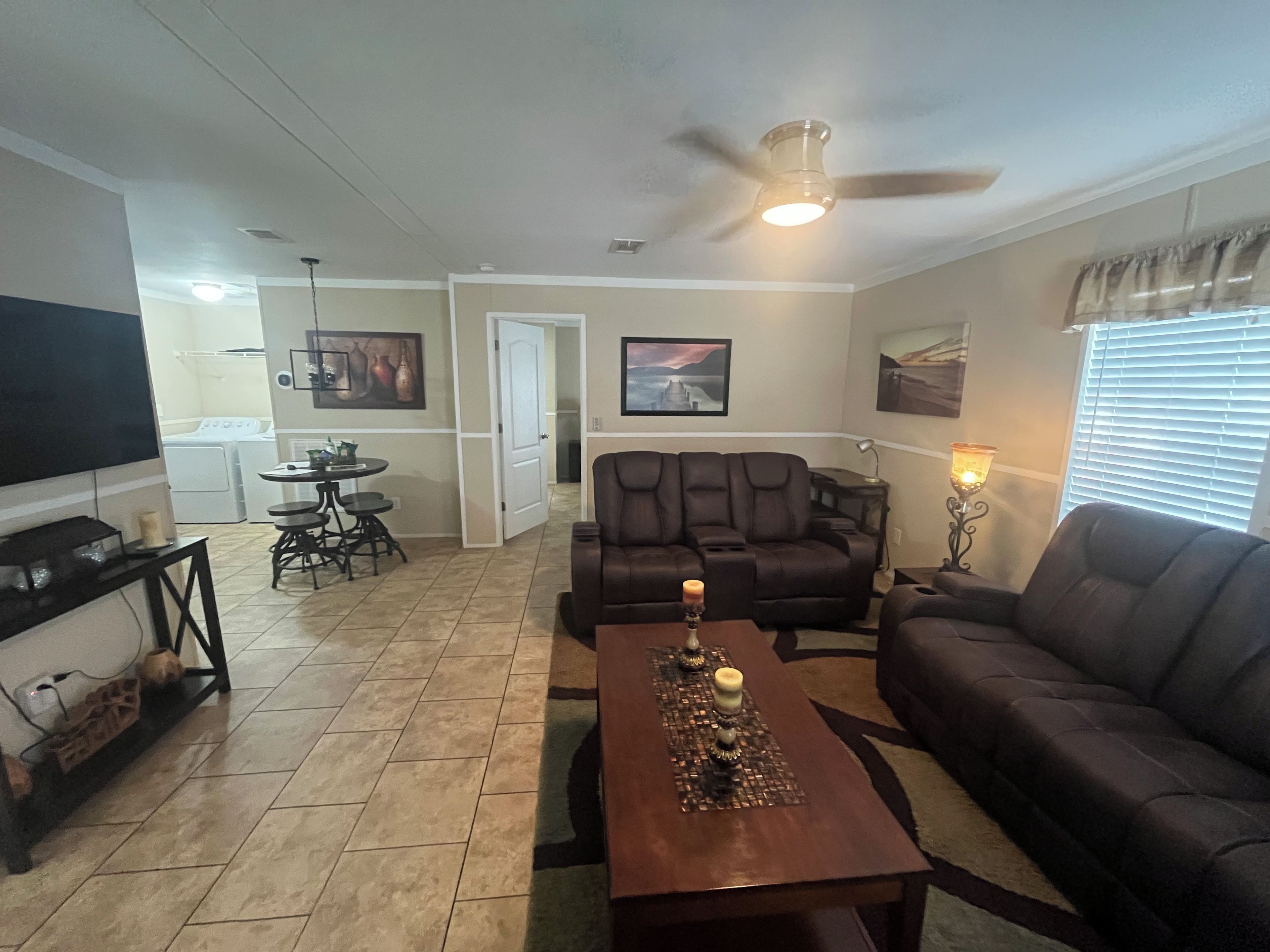 A living room with a sofa, a coffee table with candles, a television, a dining area with a table and chairs, and a ceiling fan.