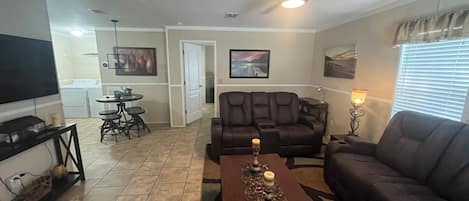 A living room with a sofa, a coffee table with candles, a television, a dining area with a table and chairs, and a ceiling fan.