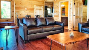 A cozy cabin interior with a leather sofa, wooden dining table, and a kitchen area visible through an open door. - Gorgeous Smoky Mountain Views! Amazing Views Near Pigeon Forge & Gatlinburg! (Sevierville)