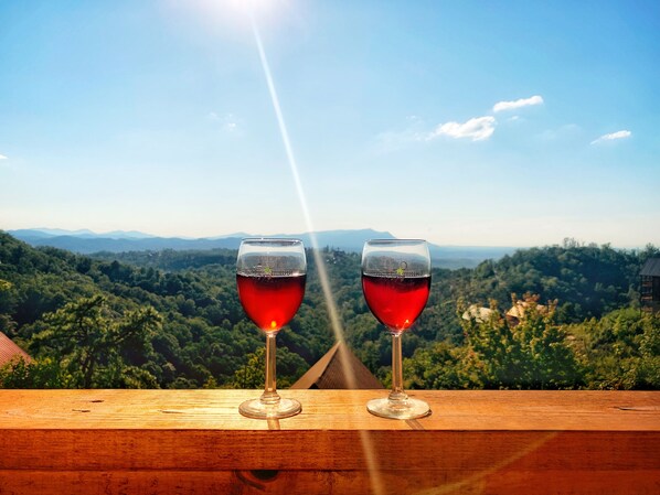 Dos copas de vino sobre un repisa de madera, con una vista panorámica de un paisaje boscoso y montañas al fondo.