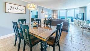 A dining area with a wooden table and chairs, a chandelier, and a 'gather' sign on the wall. - 🌅 Free Seasonal Beach Chairs • Gulf Views • Shoreline 3073 (Destin)