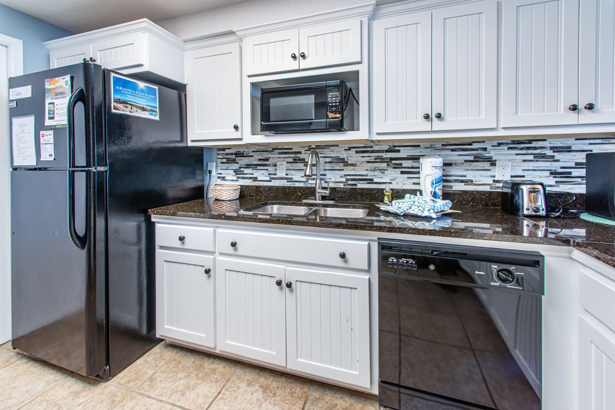 A modern kitchen with white cabinets, a stainless steel refrigerator, a microwave, a dishwasher, and a granite countertop.