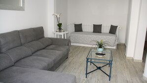 A living room with a grey sofa, a small round table with a plant, and a bed with grey bedding.