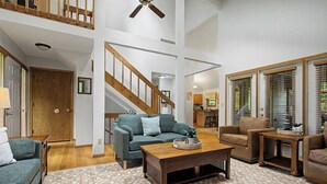 A spacious living room with a wooden coffee table, a sofa, and a chair. A staircase with wooden railings leads to an upper level.