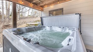 A hot tub with a wooden ceiling and string lights, surrounded by a stone wall and trees.