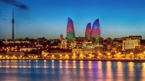 A cityscape at dusk with illuminated buildings, a prominent tower, and a waterfront. - Квартира центр города (Baku)