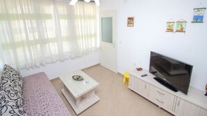 Un soggiorno con divano, un tavolino, una TV su un mobile e una finestra con tende leggere.