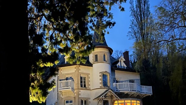 Una casa gran de moltes plantes amb una torre destacada, un balcó i un interior ben il·luminat.