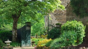 Un camí al jardí que condueix a un edifici de pedra amb una porta metàl·lica, envoltat de vegetació exuberant i diverses plantes.