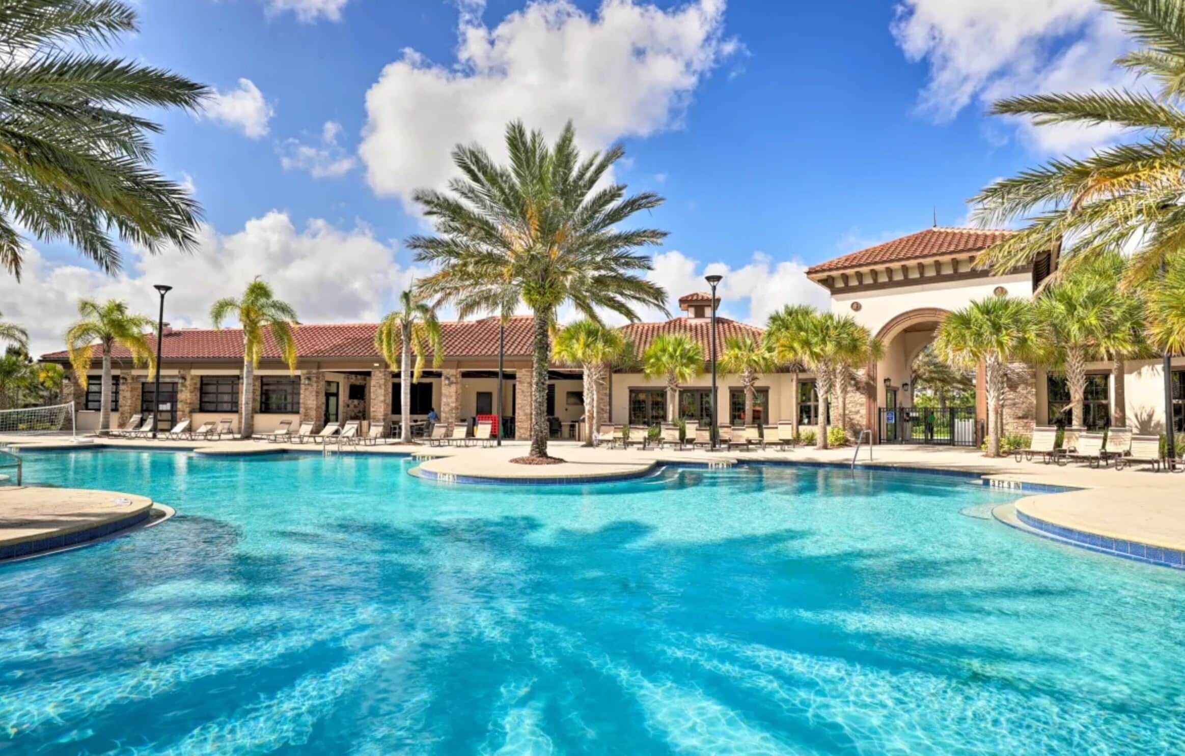A swimming pool with lounge chairs and palm trees, surrounded by a building with a tiled roof and stone walls.