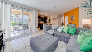 A living room with a grey sofa, ottoman, and green cushions. There is a sliding glass door leading to a patio with a table and chairs.