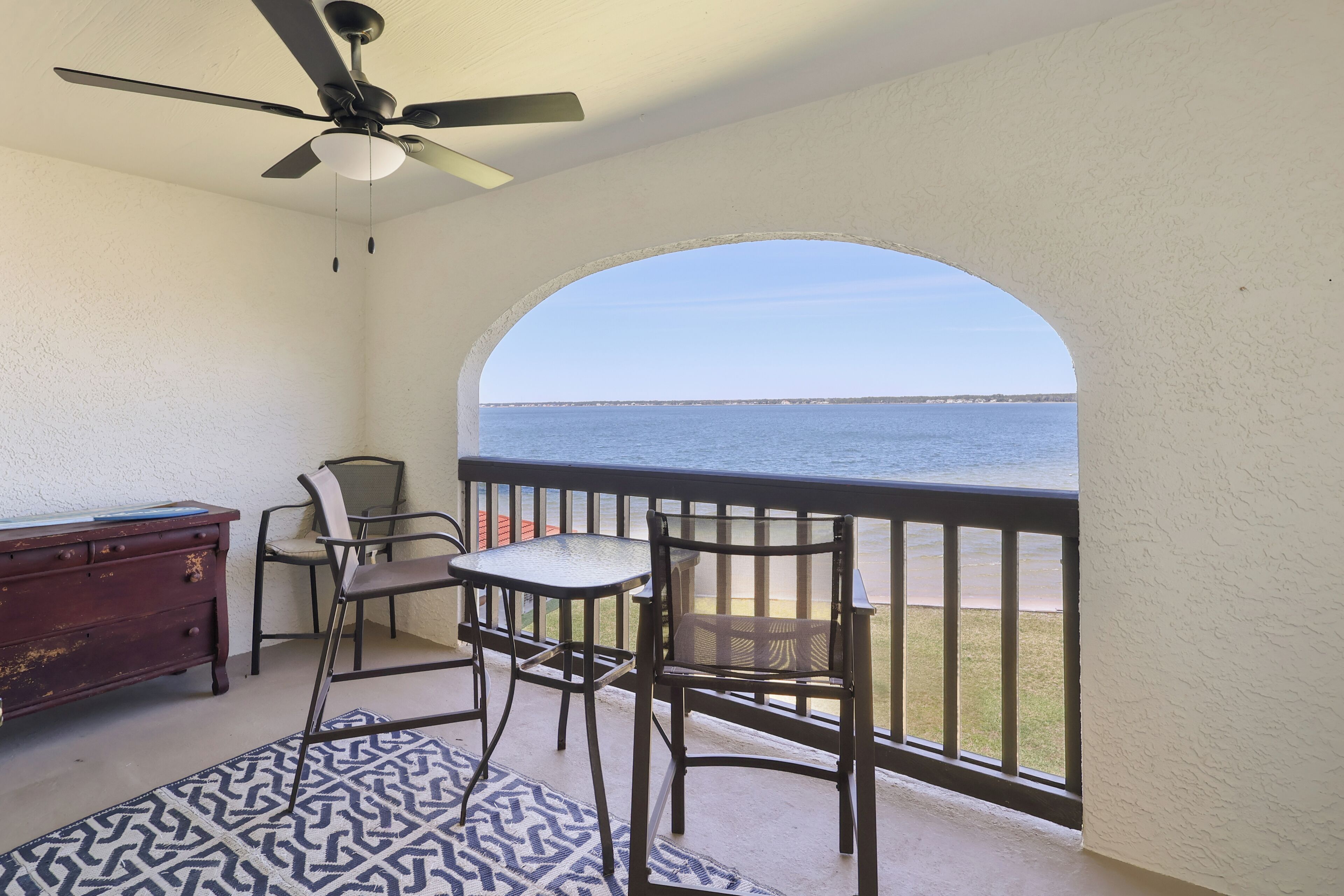 A balcony with a ceiling fan, a table, chairs, a patterned rug, and a view of the ocean.