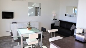 Un salon moderne avec une table à manger au plateau en verre, des chaises blanches, un canapé noir, un téléviseur fixé au mur et un petit coin cuisine équipé d’un grille-pain.